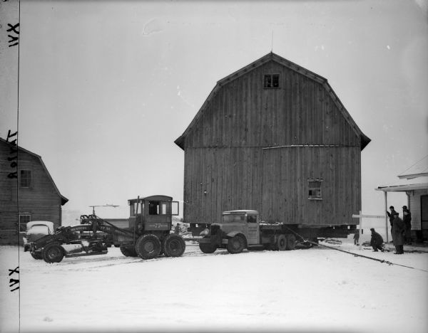 Taylor barn being moved.  A tractor and a truck are pulling the barn, which is on a platform, through the snow. A smaller barn is on the left.  On the right, five people are watching from a porch.