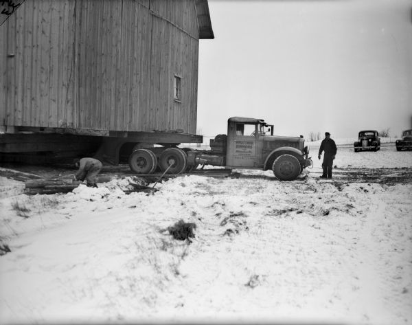 The Taylor barn is being moved by a truck in the snow. One man is standing in front of the truck, and another man is bending over on the left near a pile of wood near the barn. Two cars are in the background.