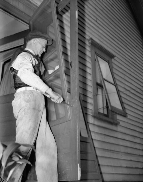 J. Robert Taylor fixing a screen door. He is standing on the inside of the door, with a hammer in his right hand.