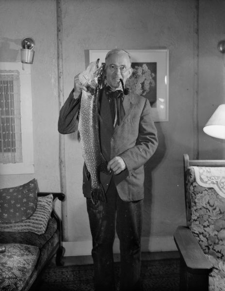 The photographer holding a large Northern Pike and smoking a pipe in a living room.