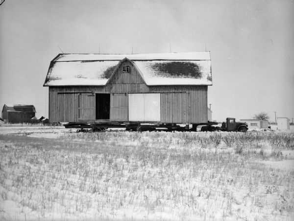 The Taylor barn being moved through snow-covered fields by a truck. The barn is on a large bed platform. Other farm buildings are in the background.