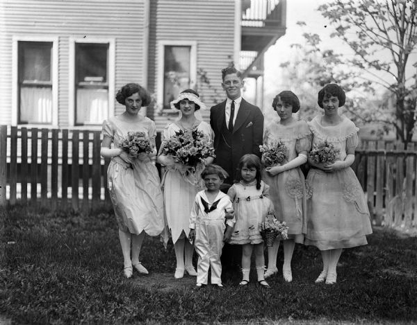 Outdoor portrait of a wedding party that includes the bride and groom (center), three bridesmaids, a flower girl, and a ring bearer. A fence and a house are in the background.