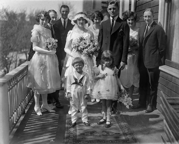 Outdoor group portrait of a wedding party on a balcony. The party consists of the bride and groom (center), three bridesmaids, three groomsmen, a flower girl, and a ring bearer. The two children are standing on a carpet.