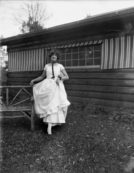 Outdoor portrait of a woman in a white dress identified as Donna Taylor Adams, J. Robert Taylor's daughter. She is standing next to a bench and is holding her skirt in her hands. A building is behind her.