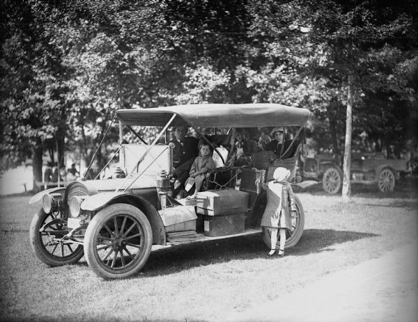 A car (possible a Model T) parked in the grass with a family sitting inside. There are other people with cars in the background under trees, and people sitting at picnic tables.