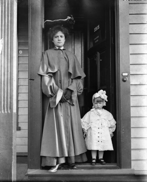 Alma Reinhardt Taylor and her toddler daughter Ellen Taylor Higgins standing in a doorway. Both are wearing long, cool weather coats. The number on the door is 3315, which stands open behind them.