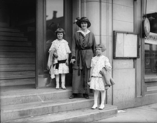 A unidentified woman is standing on a stairway outside a doorway with two young girls, identified as Donna Taylor on the right, and Ellen Taylor on the left. The young girls are carrying their coats over their arms.