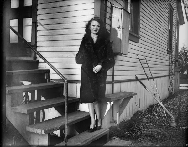 Portrait of a young woman leaning against the back corner of a house while standing on the steps. She is wearing a fur coat.