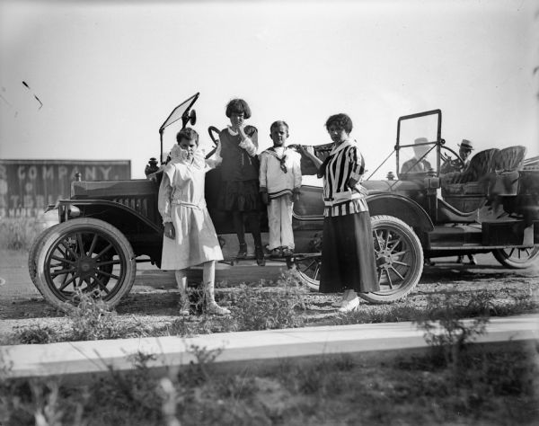 A group portrait taken next to an automobile, consisting of a woman (probably Alma Reinhardt Taylor) with her hand on the car, one boy (probably Fred Taylor) standing on the vehicle running board, and two girls (probably Ellen and Donna Taylor). Two men are talking in the background while standing behind another car. There is a billboard in the background on the left.