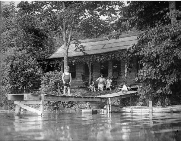 View from water of Alma Reinhardt Taylor and her three children (from left) Frederick, Donna, and Ellen outside a cabin nestled among trees along a lake. Frederick stands at the end of a pier.