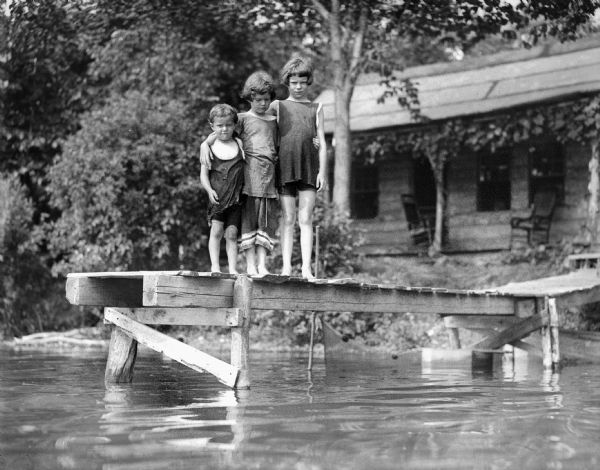View from lake towards Fred, Donna and Ellen Taylor standing on a pier. A cabin is on the shoreline in the background, nestled in trees.