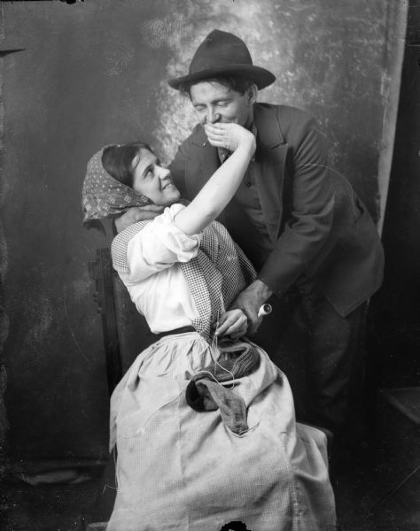 A studio portrait of Alma and J. Robert Taylor. Alma is sitting in a chair holding a sock to be darned, while putting her hand over J. Robert's mouth. He is bending over her laughing, and is holding a pipe in one hand.