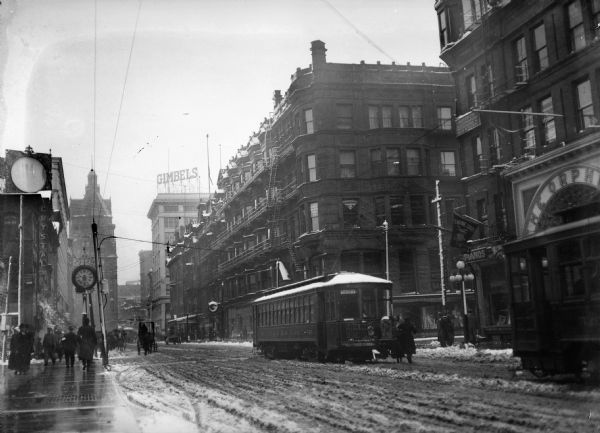 Snowy Milwaukee street with the Edgewood streetcar, pedestrians and horse-drawn carriages.  Men and women are passing by a clock on the sidewalk. A Gimbels sign is on top of a roof in the background.