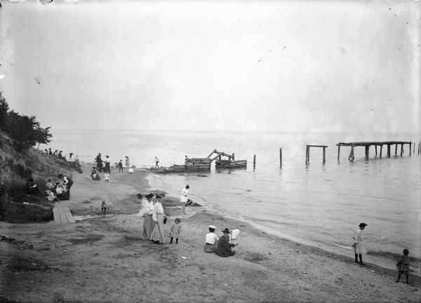 Men, women, and children sitting and walking on the shore of a lake, possibly Lake Michigan, near a partially constructed pier.