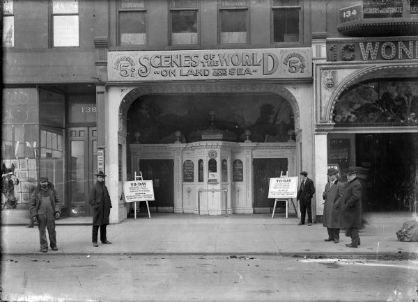 Five men are standing on a sidewalk in front of a theater. The sign above the theater reads: "Scenes of the World on Land and Sea, Admission 5 cents". The signs on easels flanking the entrance both read: "To-Day: Crushed Alive, Saved by a Melon, Married Four Millions. Songs: When the Golden Rods Were Waving Mollie Dear, The Waltz Must Change to a March Marie".