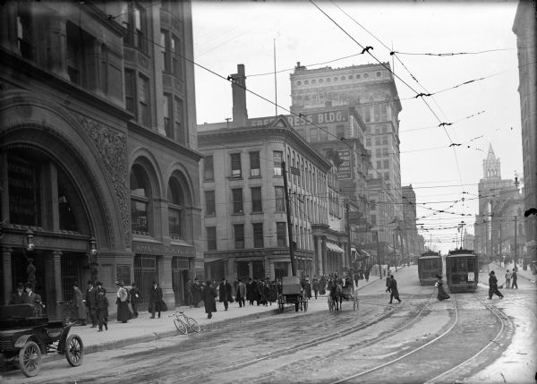 Street and sidewalk outside of the Wisconsin National Bank. Men, women, and children are walking on sidewalks, and crossing in front of streetcars. Horse-drawn carriages, a bicycle, and an automobile are also moving along the street.