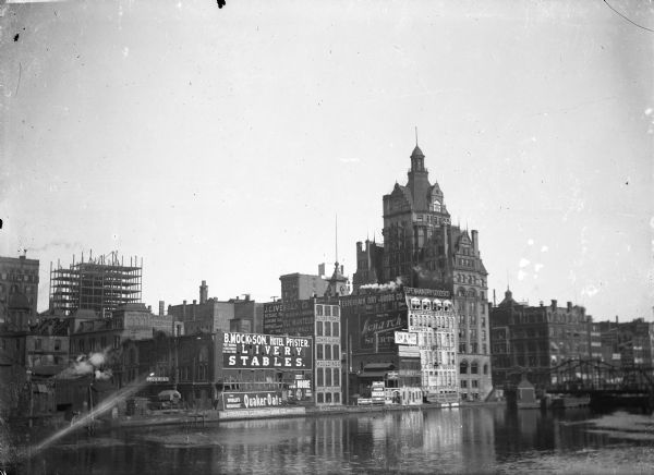 View of downtown Milwaukee riverbank before the First National Bank was built in 1914. Advertising painted on the sides of buildings includes  B. Mock & Son, Hotel Pfister Livery Stables, Quaker Oats, J.C. Iversen Co., Espenhain Dry Good Co., and Monarch Shirts. There is a bridge over the river on the right side.