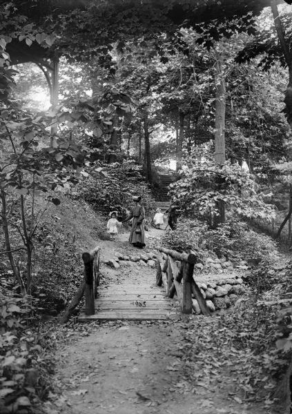 A woman and young girl in a bonnet, possibly mother and daughter, and a man and a young girl, possibly father and daughter, holding hands while strolling on a path near a wooden bridge in Lake Park.