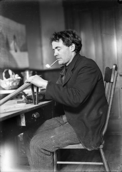 Portrait of the photographer J. Robert Taylor sitting at a table smoking a pipe and possibly woodworking.