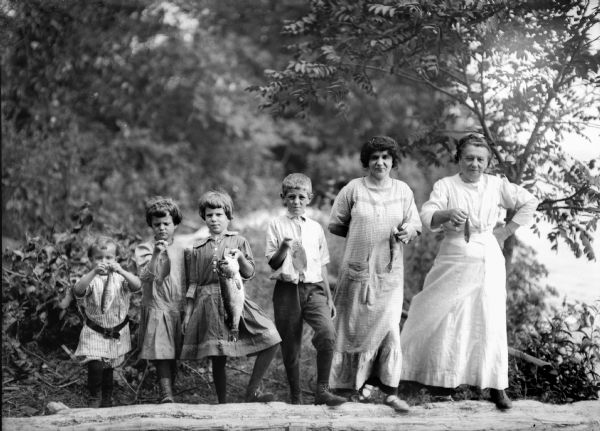 Family lined up and holding fish on what appears to be a lake shore.  From left: Fred Taylor, Donna Taylor Adams, Ellen Taylor Higgins, unidentified boy, Taylor's wife Alma Reinhardt Taylor, and Taylor's mother-in-law Katie Schaus Reinhardt.