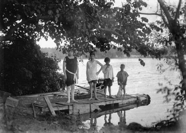J. Robert Taylor's wife, Alma Reinhardt Taylor, and children, Donna Taylor Adams, Ellen Taylor Higgins, and Fred Taylor, posed in bathing suits on a small pier.  A boat with oars and motor is visible in the background.
