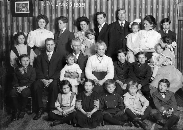 Large family group posed in parlor.  Appearing in the back row, third from the left, Alma Reinhardt Taylor holding Ellen Taylor Higgins next to  J. Robert Taylor.