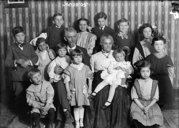 Large family group of children posed around what appears to be their grandparents in a parlor.  Standing in the middle, front row is Ellen Taylor Higgins.