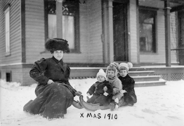 Winter scene with Alma Reinhardt Taylor and her children (from left) Frederick Taylor, Donna Taylor Adams, and Ellen Taylor Higgins.  The children are posing on a sled during a snowstorm in front of a house, Milwaukee, Wisconsin.
