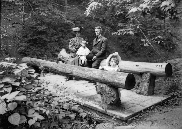 The J. Robert Taylor family on a log pedestrian bridge in a wooded area.  Identified as Alma Reinhardt Taylor, Donna Taylor Adams, J. Robert Taylor, and Ellen Taylor Higgins.