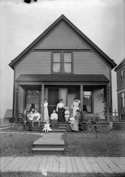 Family posed on the front porch of a home.  A wooden sidewalk appears in the foreground of the photograph.  J. Robert Taylor appears at the far left of the image.