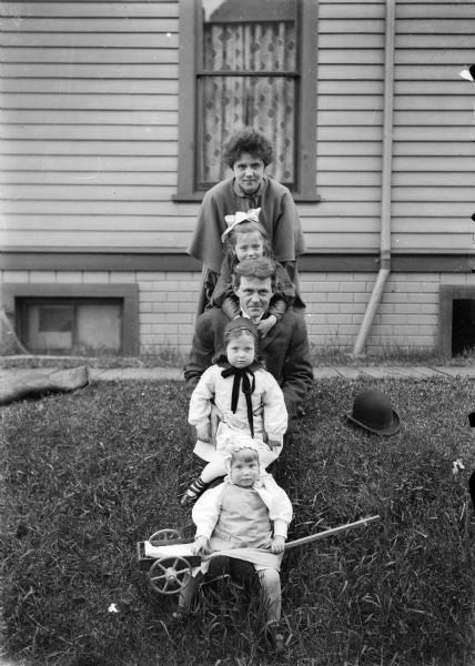 Family posed in a line in a yard outside a home.  Identified from back: Alma Reinhardt Taylor, Ellen Taylor Higgins, J. Robert Taylor, Donna Taylor Adams, and Fred Taylor sitting on a small wagon.  A bowler hat sits on the lawn near the family.