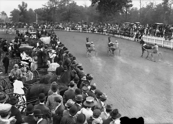 Elevated view of a horse racing track and spectators.  Sulkies, which are two-wheeled, one-person carriages drawn by a single horse, are racing.  The race possibly took place at Washington Park.