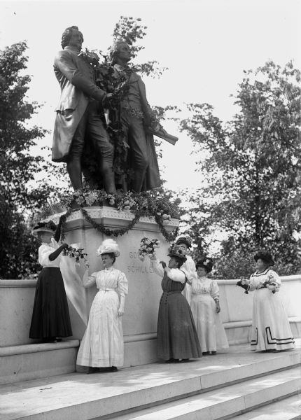 Women placing floral garlands on a monument to Johann Wolfgang von Goethe and Friedrich von Schiller.  The women are wearing decorative hats.
