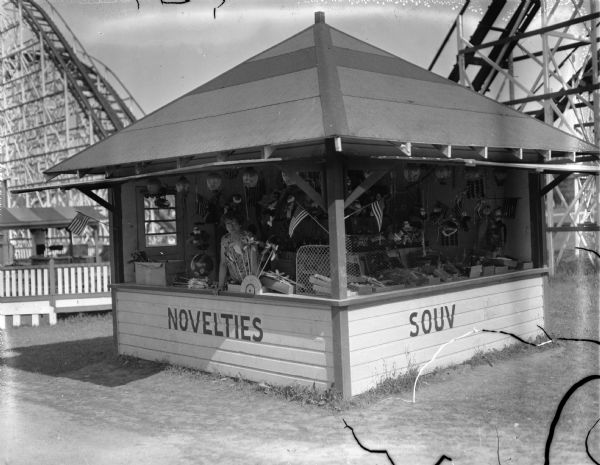 A woman staffing a souvenir booth at a fair. Souvenirs being sold include pinwheels and flags.  The booth has "NOVELTIES" and "SOUV" painted on its sides. A roller coaster is in the background.