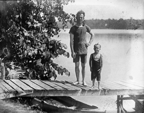 J. Robert Taylor standing on a lake pier with either his son Frederick or Robert. Both of them are wearing swimming suits, and a boat is underneath the pier at the shoreline. Taylor is smoking a pipe.