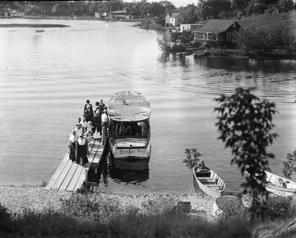 Elevated view from hill of men and women disembarking from the "Yankee Boy" boat on what appears to be a lake pier, with rowboats on the near shoreline, and homes on the far shoreline in the background.
