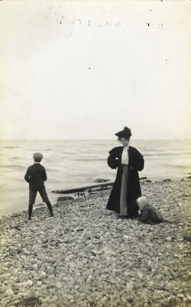 A woman, possibly Alma Reinhardt Taylor, wearing a long coat, on a rocky beach with a boy looking out at the water, and a younger child crouched at her feet. The body of water is probably Lake Michigan.