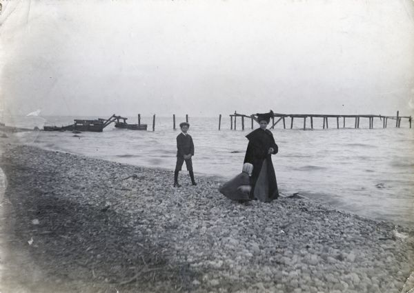 A woman, possibly Alma Reinhardt Taylor, wearing a long coat, and a young boy and toddler walking along the rocky shoreline of what is probably Lake Michigan.  A partially constructed wooden pier or pilings appear in the background.