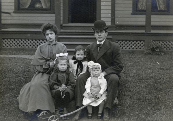 J. Robert Taylor and his wife, Alma Reinhardt Taylor, photographed in the front yard of a home with three young girls. The youngest girl is holding the handle of a two-wheeled toy wagon.