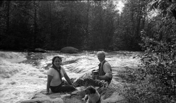 J. Robert Taylor, his niece Fern Stauff Peters, and a dog are sitting on rocks near a river. Taylor is holding what appears to be a camera.