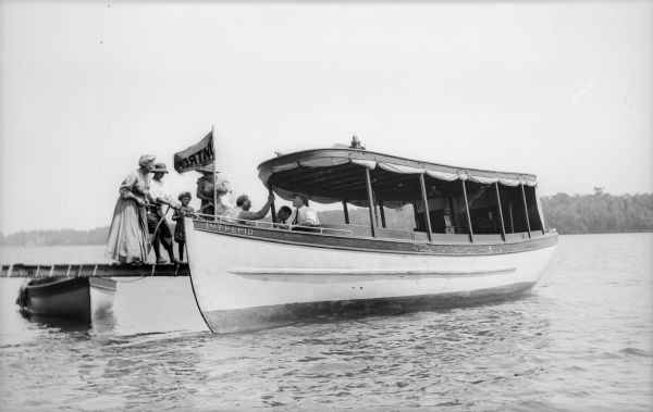 View across water towards a group of people on a pier with the boat <i>Intrepid</i>. One child is holding a rope connected to the boat, which is being driven by a man wearing a hat and tie. The boat has a flag that reads <i>Intrepid</I>. There is a smaller boat on the left.