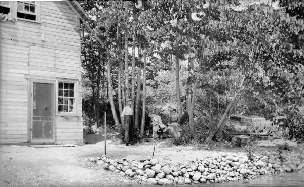 Members of the J. Robert Taylor family. A man is standing near two women who are sitting under a grove of trees near the rocky shoreline of a body of water. One of the woman is holding her hand above a dog who is sitting on its hind legs waiting for a treat. To the left of the group is what appears to be a cabin with a screen door. To the right is a hammock strung from two trees.