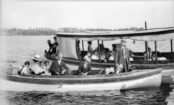 Men, women, and children aboard two boats positioned side by side in an unidentified body of water. The canopied boat in back is the "Intrepid". A man and a dog are standing at the front of the smaller boat.
