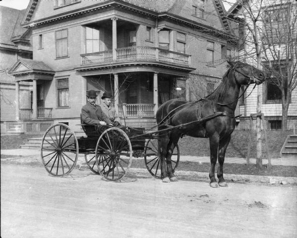 Two men wearing hats and coats in a horse-drawn carriage on a residential street.