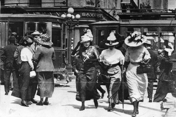 Three women in large hats walking away from two streetcars on a windy day. Other men and women are standing in the background. One of the streetcars has a sign that reads: "Delaware". A brick building on a corner behind the streetcars has a sign that reads: "Savings".