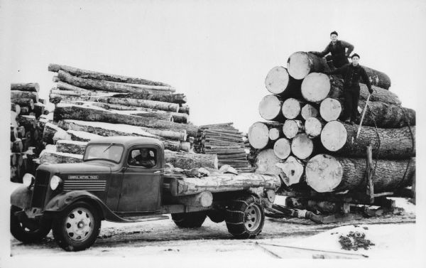 A man driving a small General Motors truck, with chains on its rear wheels, is pulling a trailer on sleds carrying large logs. Two men are standing on the trailer. Large piles of logs are visible in the background.