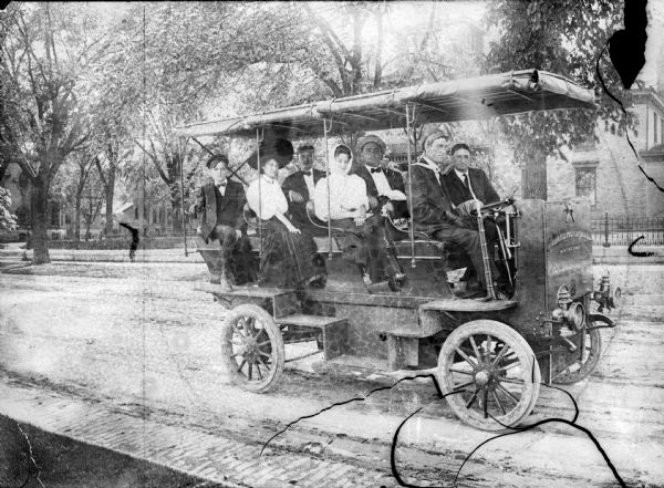 A group of two women and four men and one boy are sitting in an omnibus parked on a residential street. The front of the vehicle has a sign that appears to read: "Idaho Automobile".