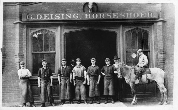Nine men in aprons lined up in front of a brick horseshoe shop. One of the men is sitting on a horse. The sign above the door reads: "C. Deising, Horseshoer," with a horse's head sculpture mounted above.