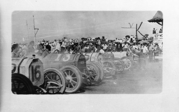 A line of cars ready for the start of a race, possibly taking place at the State Fair. Spectators are in the background.
