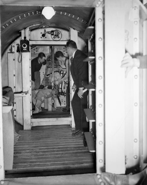 John Craig, Max Gene Nohl (seated in the dive suit), Dr. Edgar End and a fourth unknown person looking in. The location is the Milwaukee County Emergency Hospital in Milwaukee inside the decompression chamber, probably in 1937. Max was using his new self-contained dive suit and testing a new helium-oxygen mixture that Dr. End devised. As a result of these experiments, along with newly devised self-contained dive gear, Max Nohl was able to set a new record deep dive in Lake Michigan on December 1, 1937.